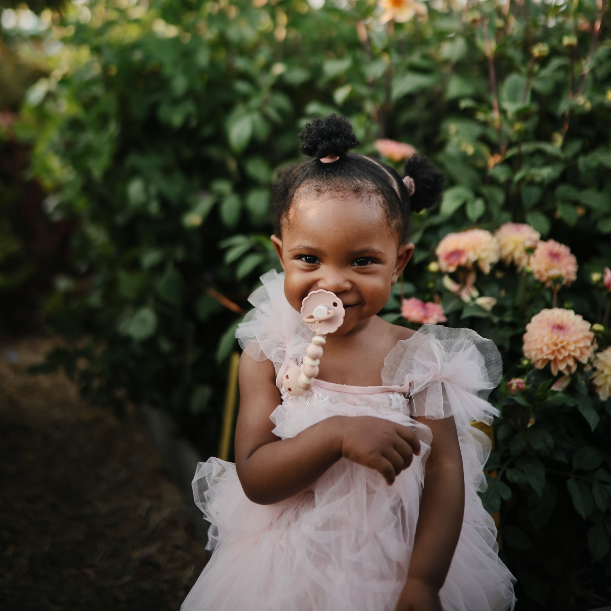 A young girl smiles in the garden, holding a FRIGG Daisy Natural Rubber Pacifier.