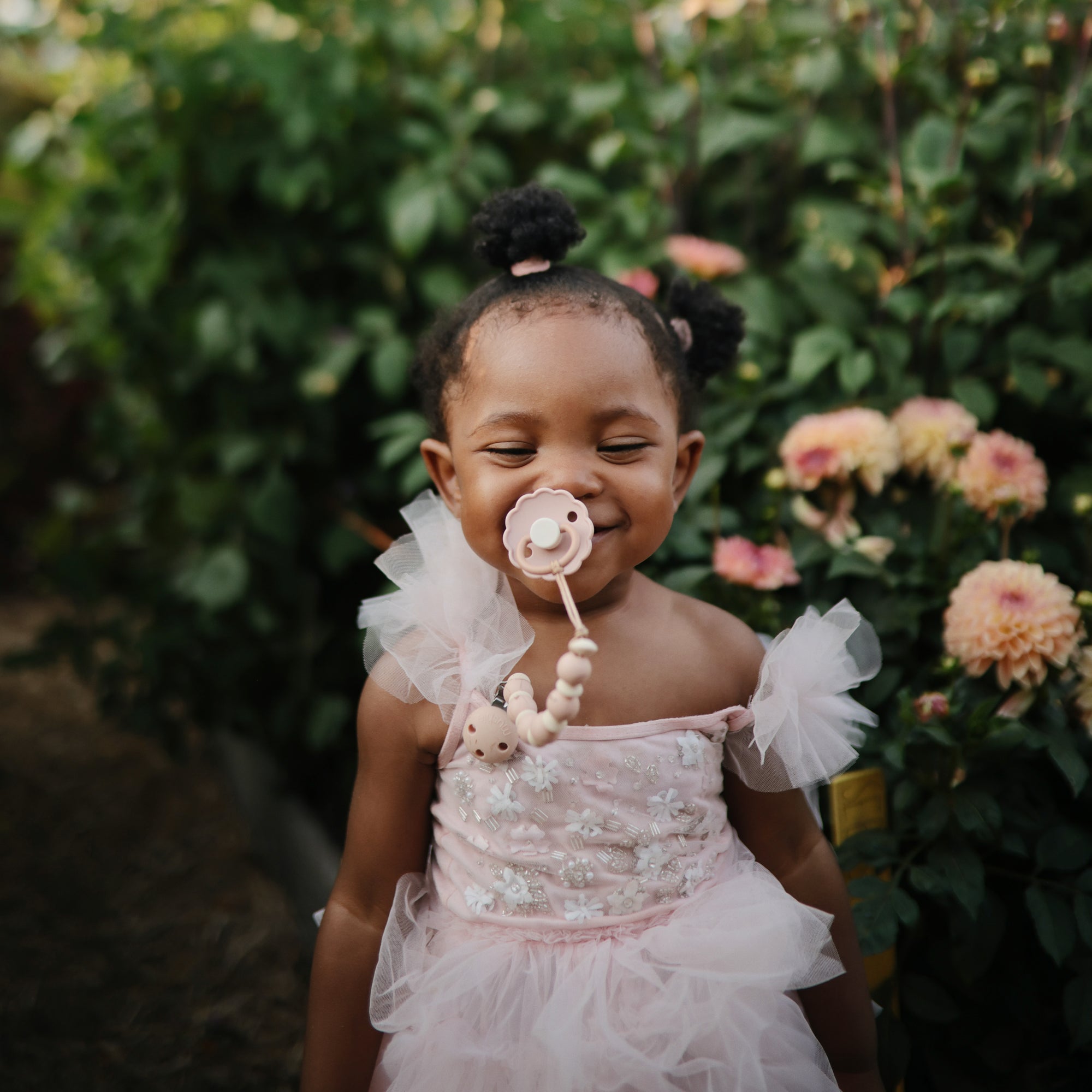 A young girl in a pink, frilly dress stands outdoors among blooming flowers, smiling with her eyes closed and using the FRIGG Daisy Silicone Pacifier 2-Pack | Limited Edition by FRIGG.