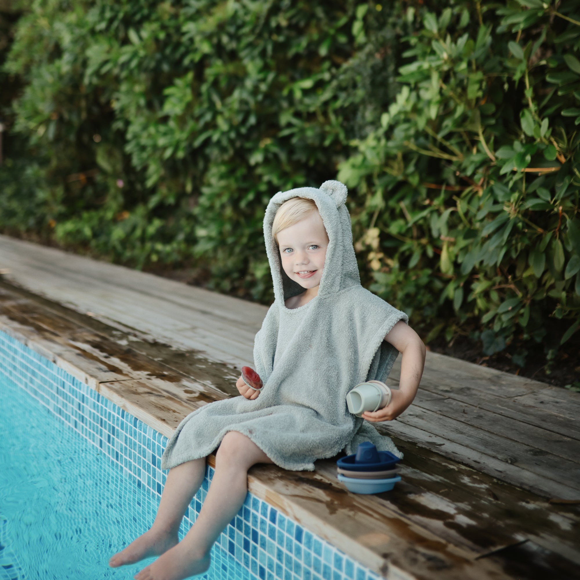 A smiling child in a mushie gray hooded towel plays with Bath Boats by the pool.