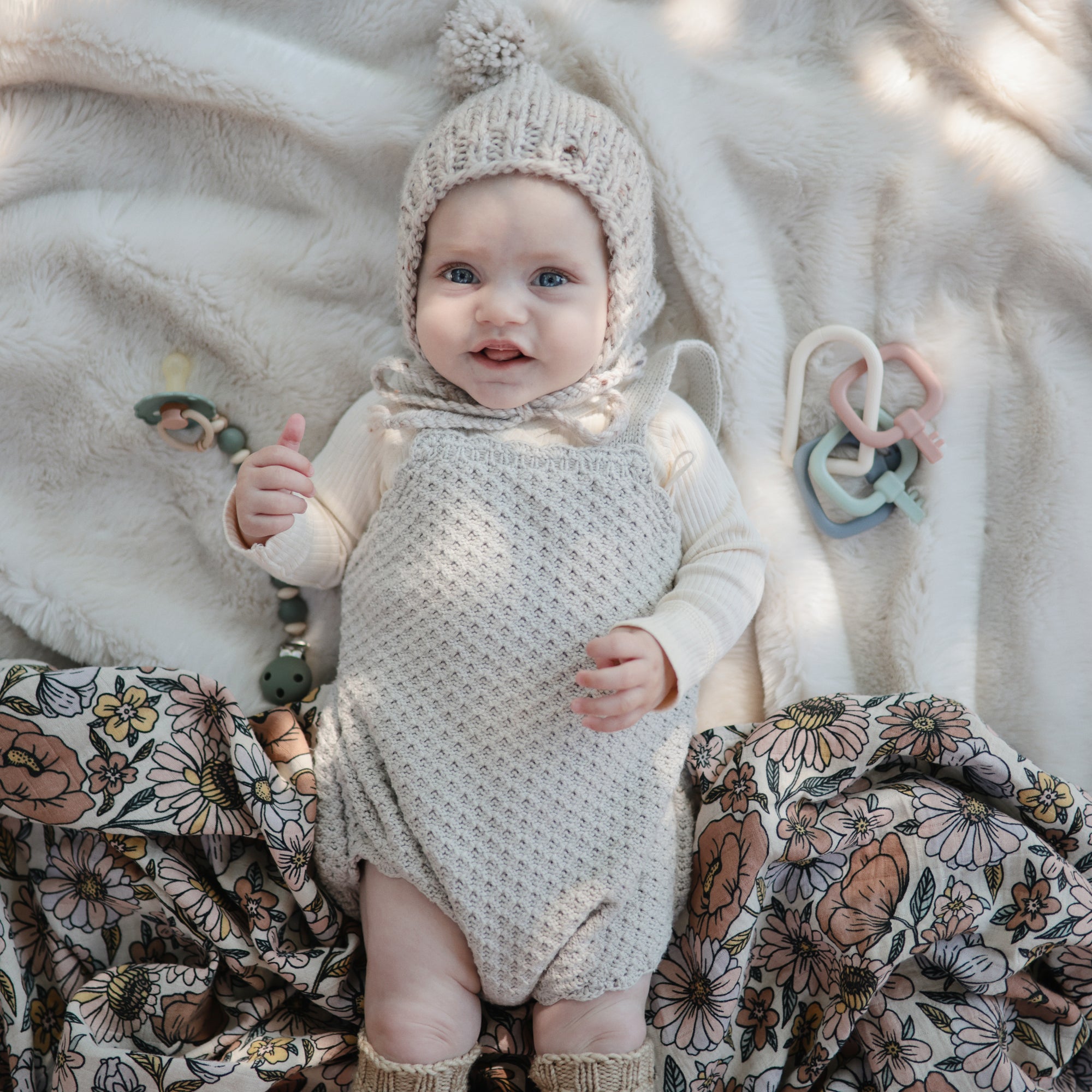 Baby on mushie Organic Cotton Muslin Swaddle Blanket, wearing knit outfit and hat with toys nearby.