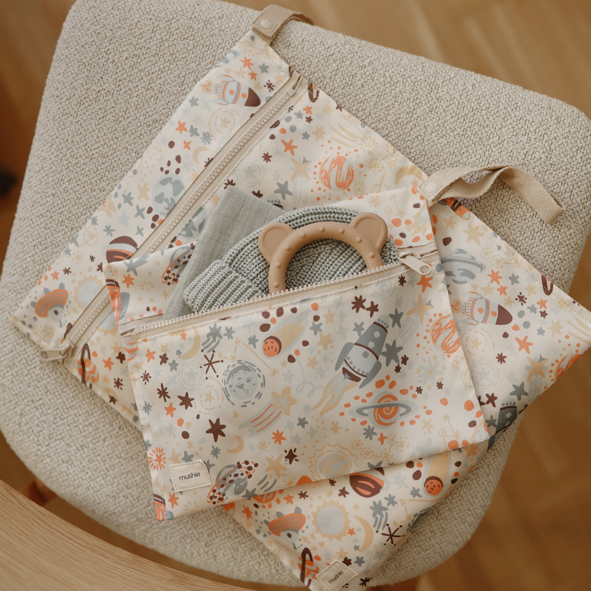 A space-patterned fabric pouch with zippers rests on a beige chair. Inside are a folded cloth and the mushie Bear Teether, a non-toxic accessory ideal for soothing little gums.