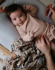 Baby in pink on pillow with mushie Organic Cotton Muslin Swaddle Blanket and floral blanket nearby.