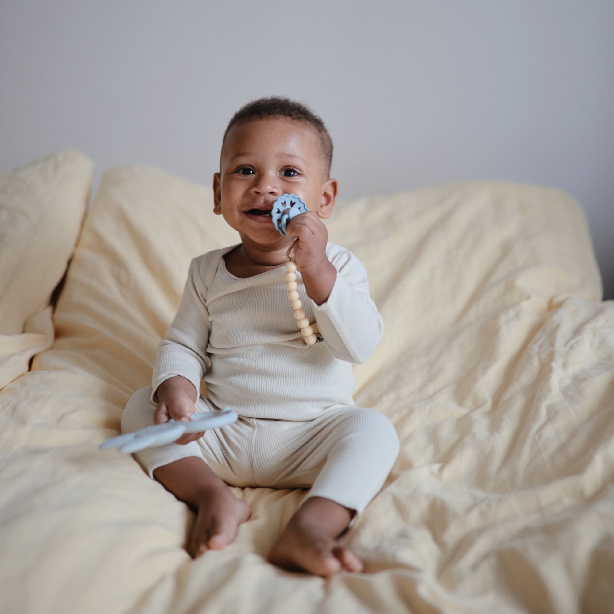 A smiling baby in cream pajamas sits on a bed with light yellow bedding, contentedly holding the FRIGG Andersen Fairytale Silicone Pacifier 2-Pack by FRIGG, made in Denmark, and a teething toy, looking playful.