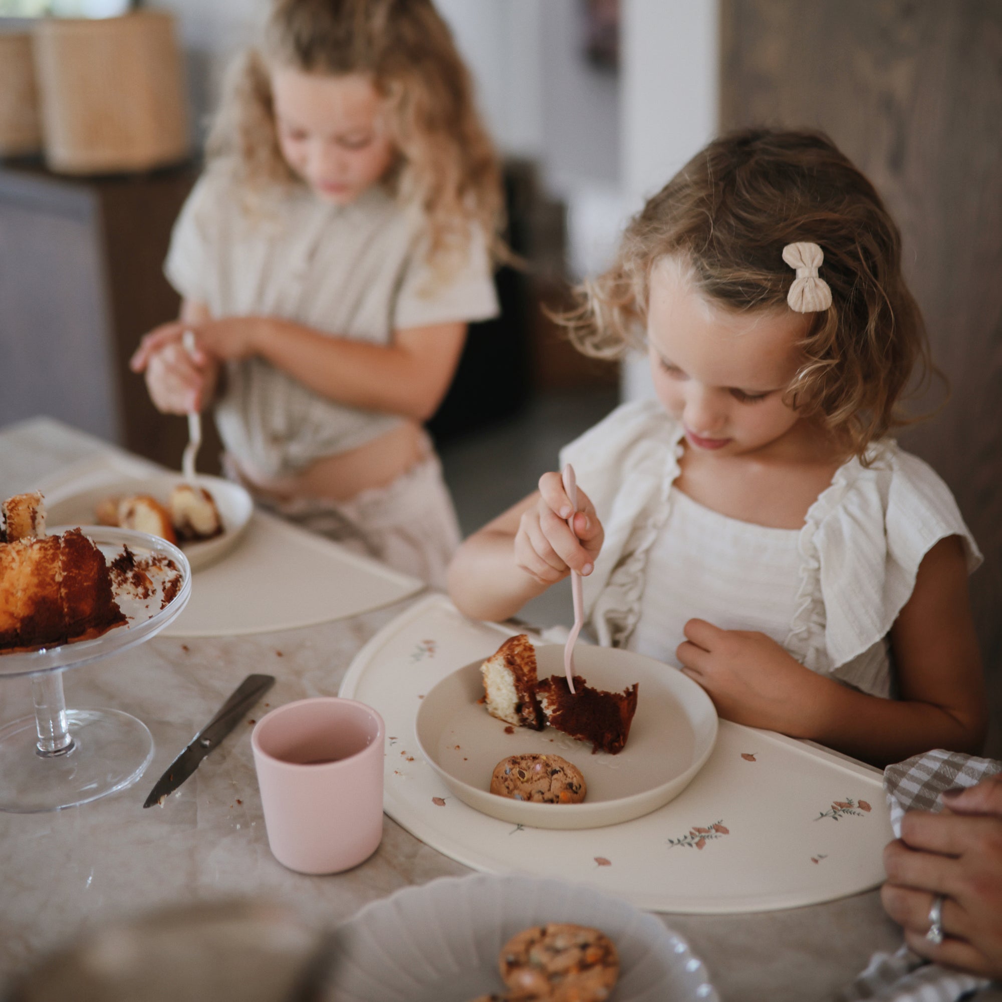 Two girls eat cake with mushie pink Dinnerware Cups and plates at the table.