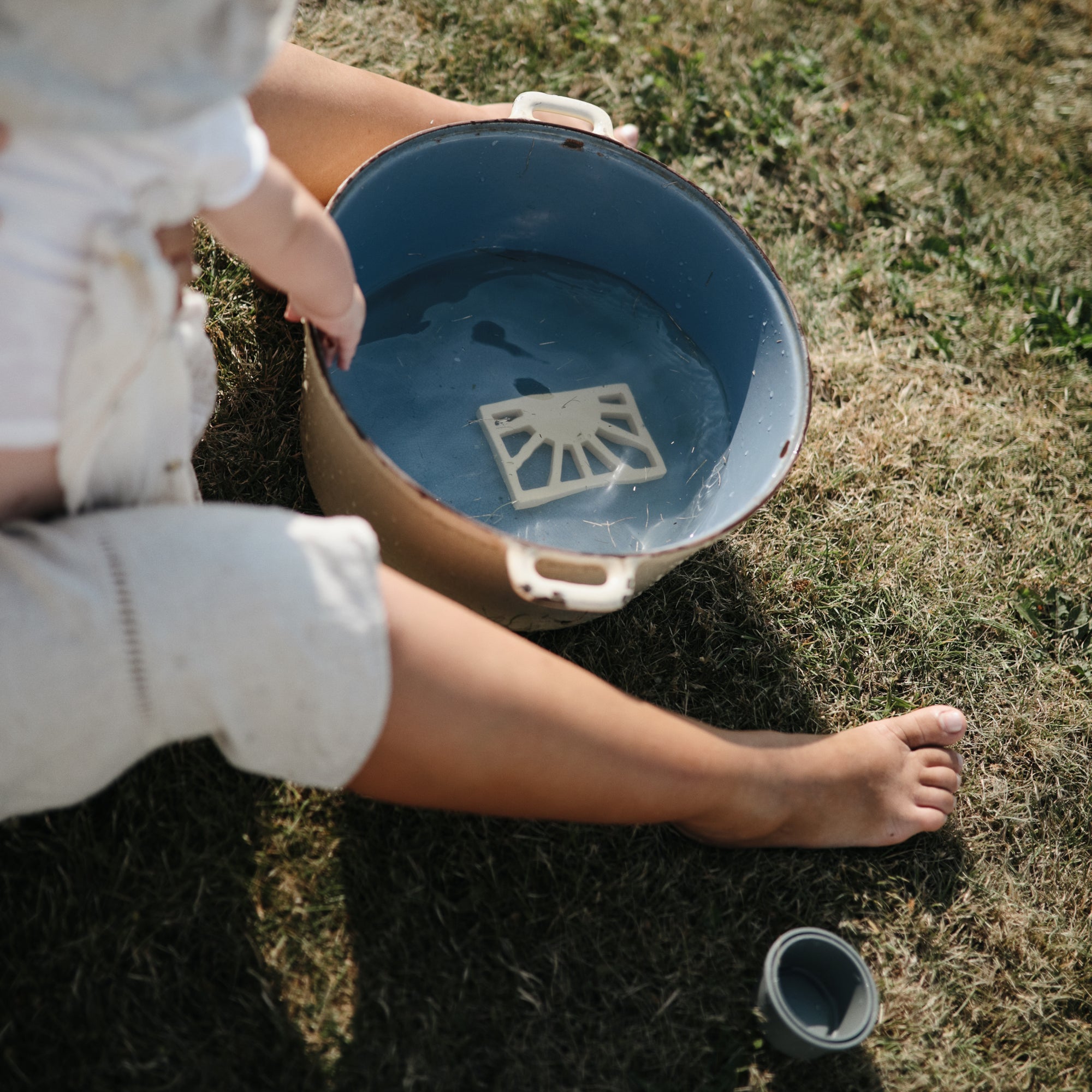 A child sits barefoot on grass next to a large blue pot of water, holding the mushie Sun Teether. A round container lid rests on the ground nearby.