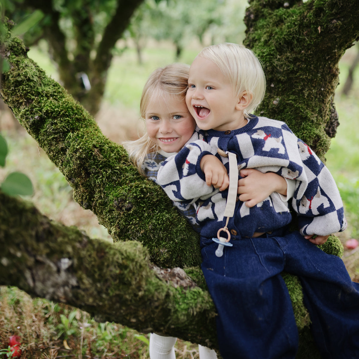 Two smiling kids, one holding mushie’s Linen Pacifier Clip 2-Pack with silicone clasp.