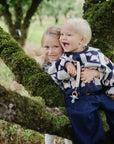 Two smiling kids, one holding mushie’s Linen Pacifier Clip 2-Pack with silicone clasp.