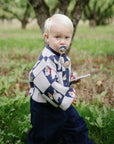 Toddler with mushie Linen Pacifier Clip 2-Pack holds a notepad in a grassy orchard, wearing sweater.