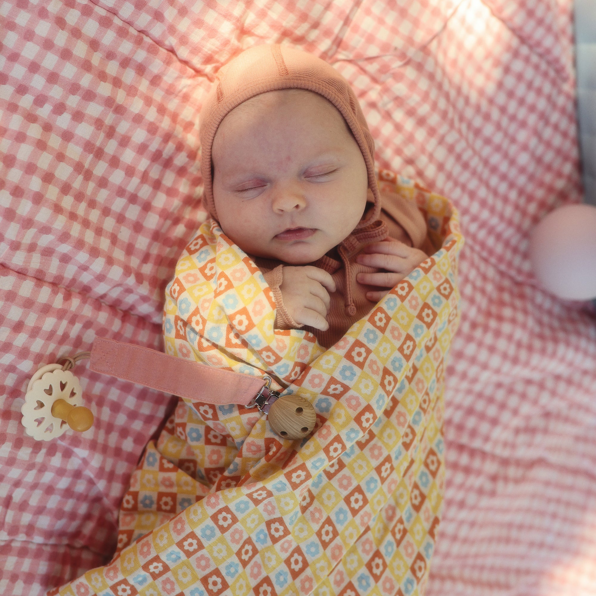 Baby in a pink hat with mushie Linen Pacifier Clip 2-Pack, sleeping on pink checks.