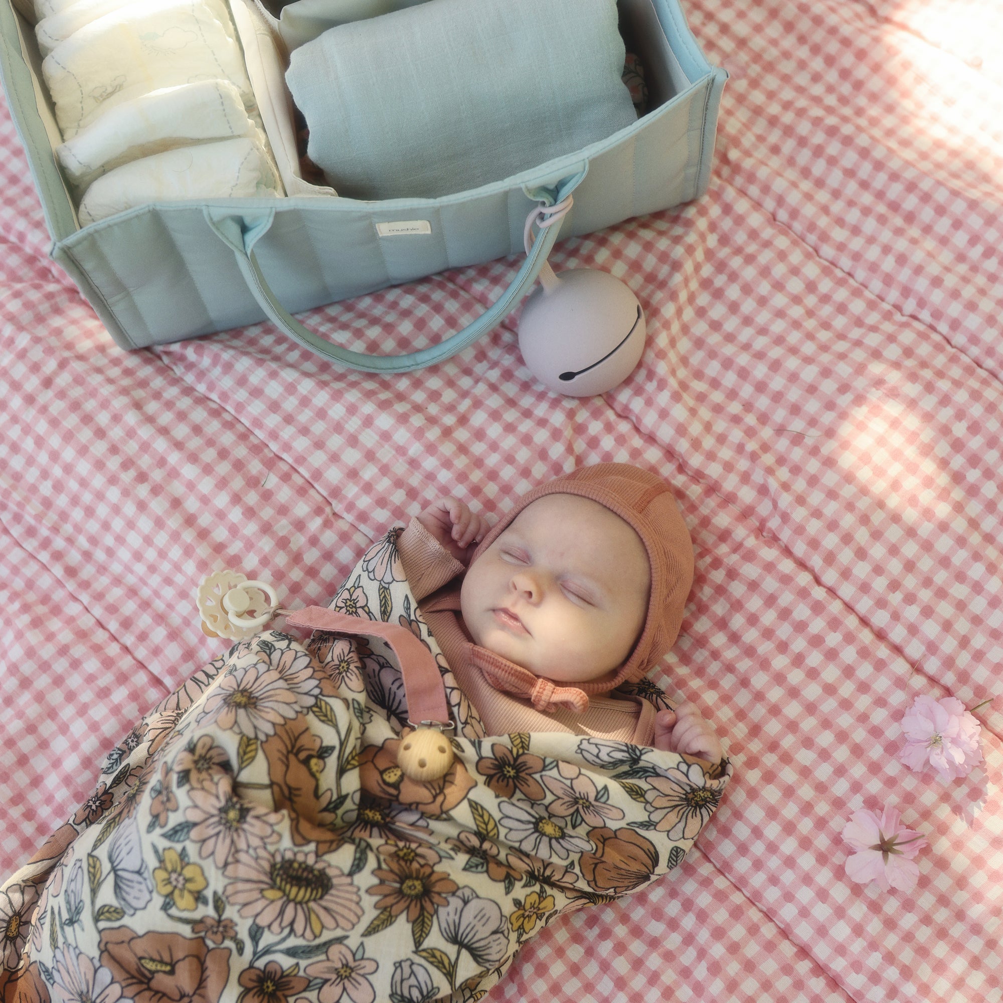 Baby sleeps on pink checkered blanket with mushie Organic Cotton Muslin Swaddle Blanket nearby.