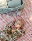 Baby sleeps on pink checkered blanket with mushie Organic Cotton Muslin Swaddle Blanket nearby.