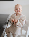 A baby in a striped bib holds a mushie Figurine Teether on a white chair by a dark table.