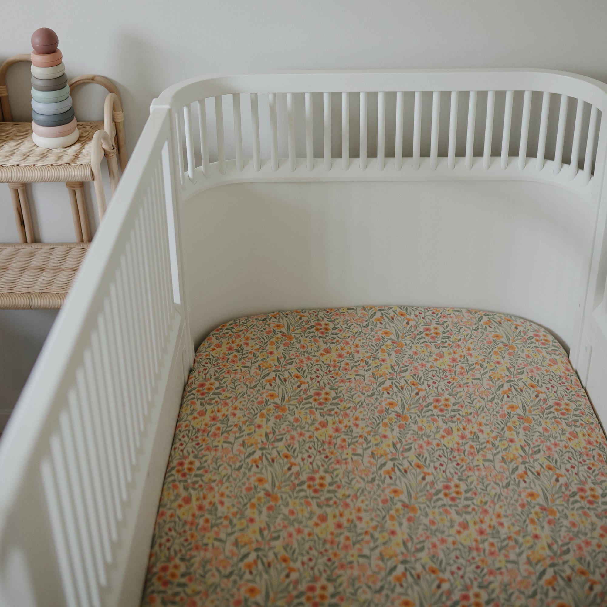 White crib with mushie Extra Soft Muslin Crib Sheet, next to a wicker shelf with pastel rings.