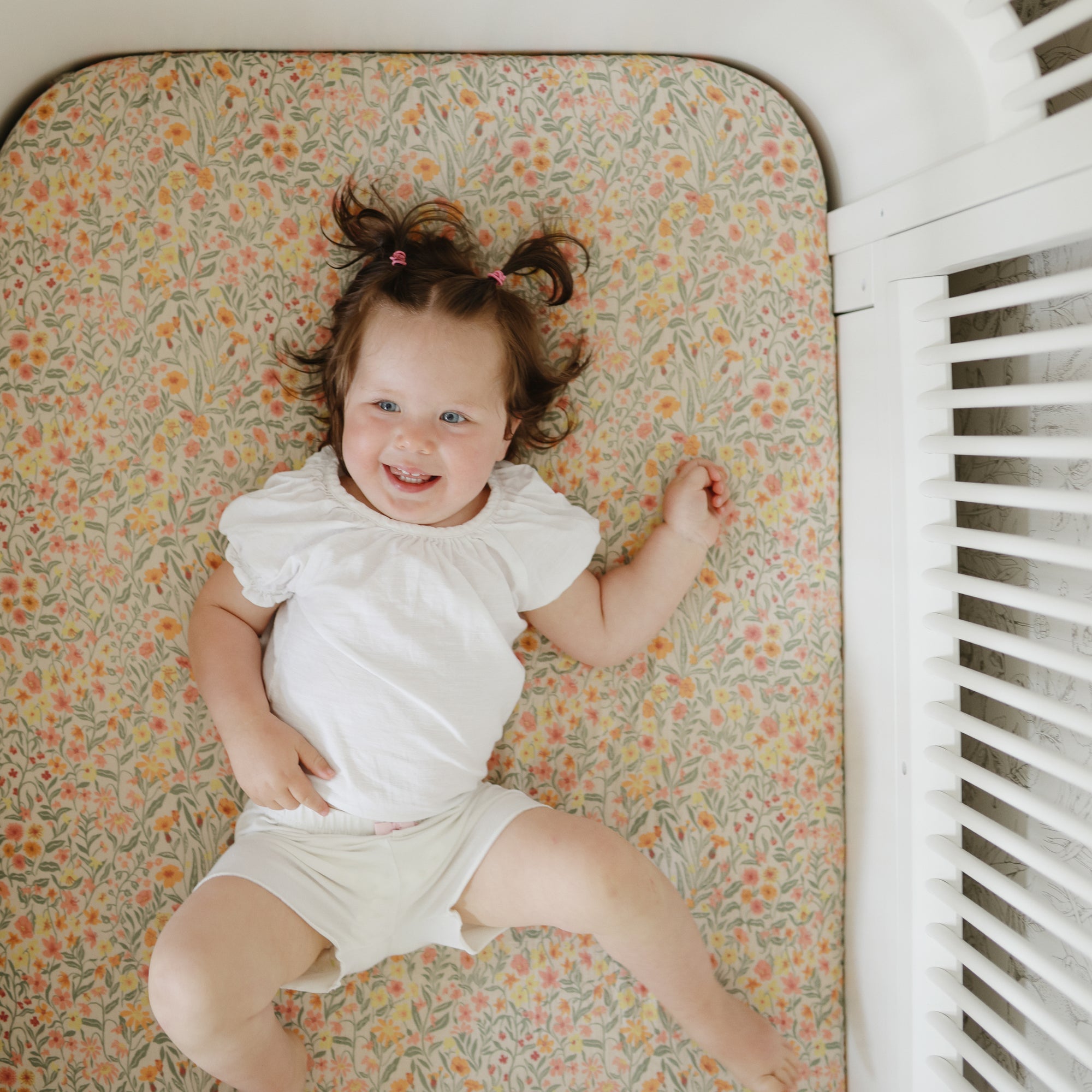Baby smiles on mushie Extra Soft Muslin Crib Sheet in white crib with floral mattress.