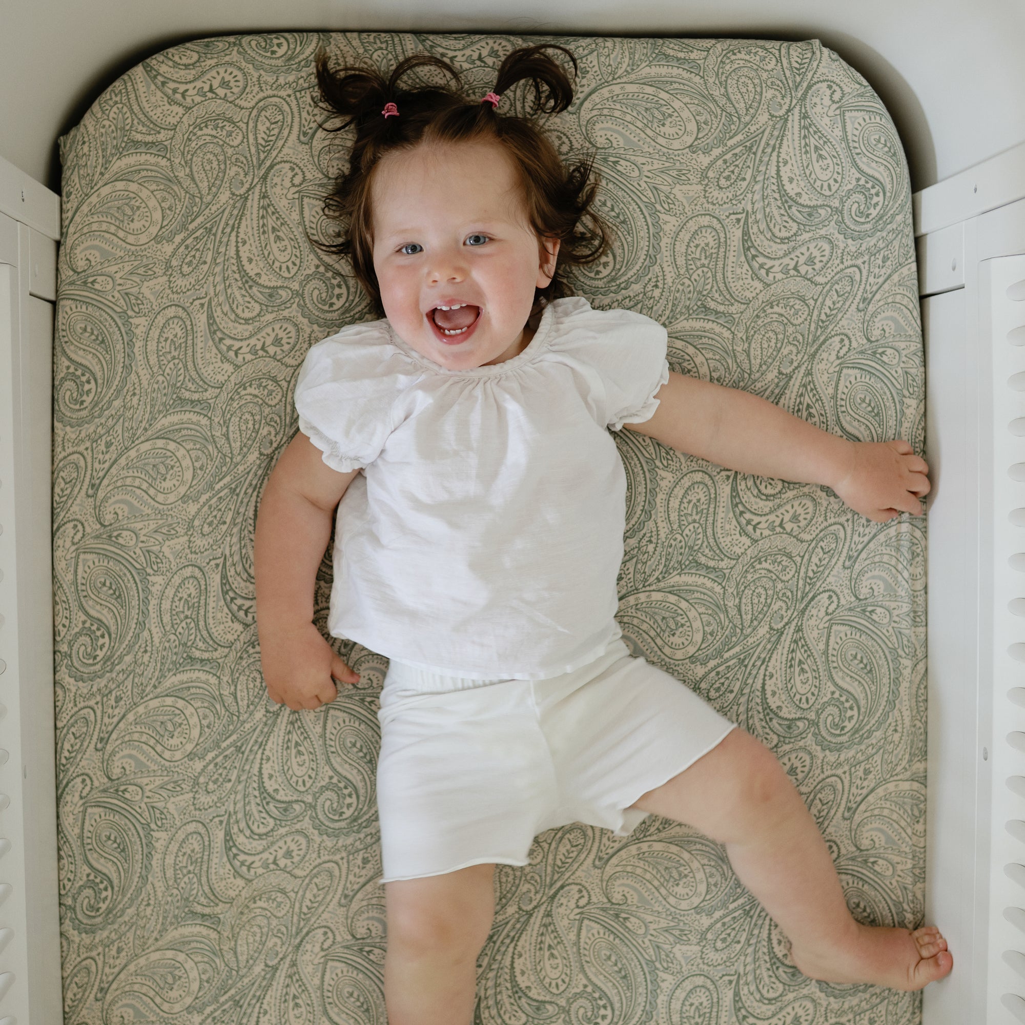 Smiling toddler on mushie Extra Soft Muslin Crib Sheet, wearing white clothes.