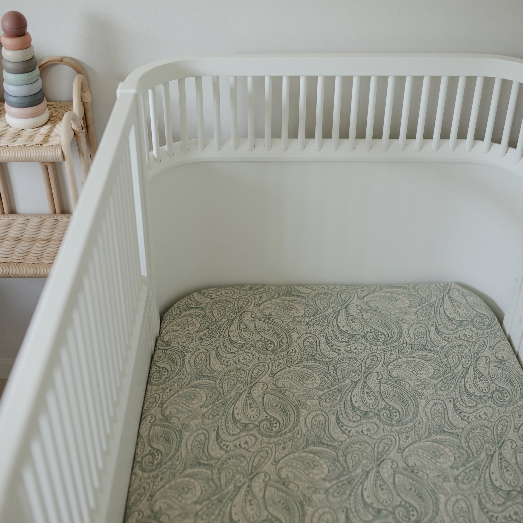Mushie Extra Soft Muslin Crib Sheet on white crib next to wicker shelf and stacked rings toy.