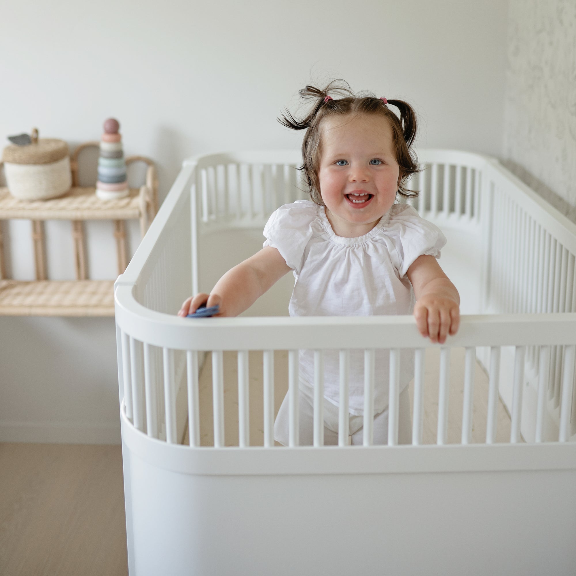 Smiling toddler stands in a mushie Extra Soft Muslin Crib Sheet, white crib, bright nursery.