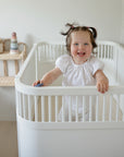Smiling toddler stands in a mushie Extra Soft Muslin Crib Sheet, white crib, bright nursery.