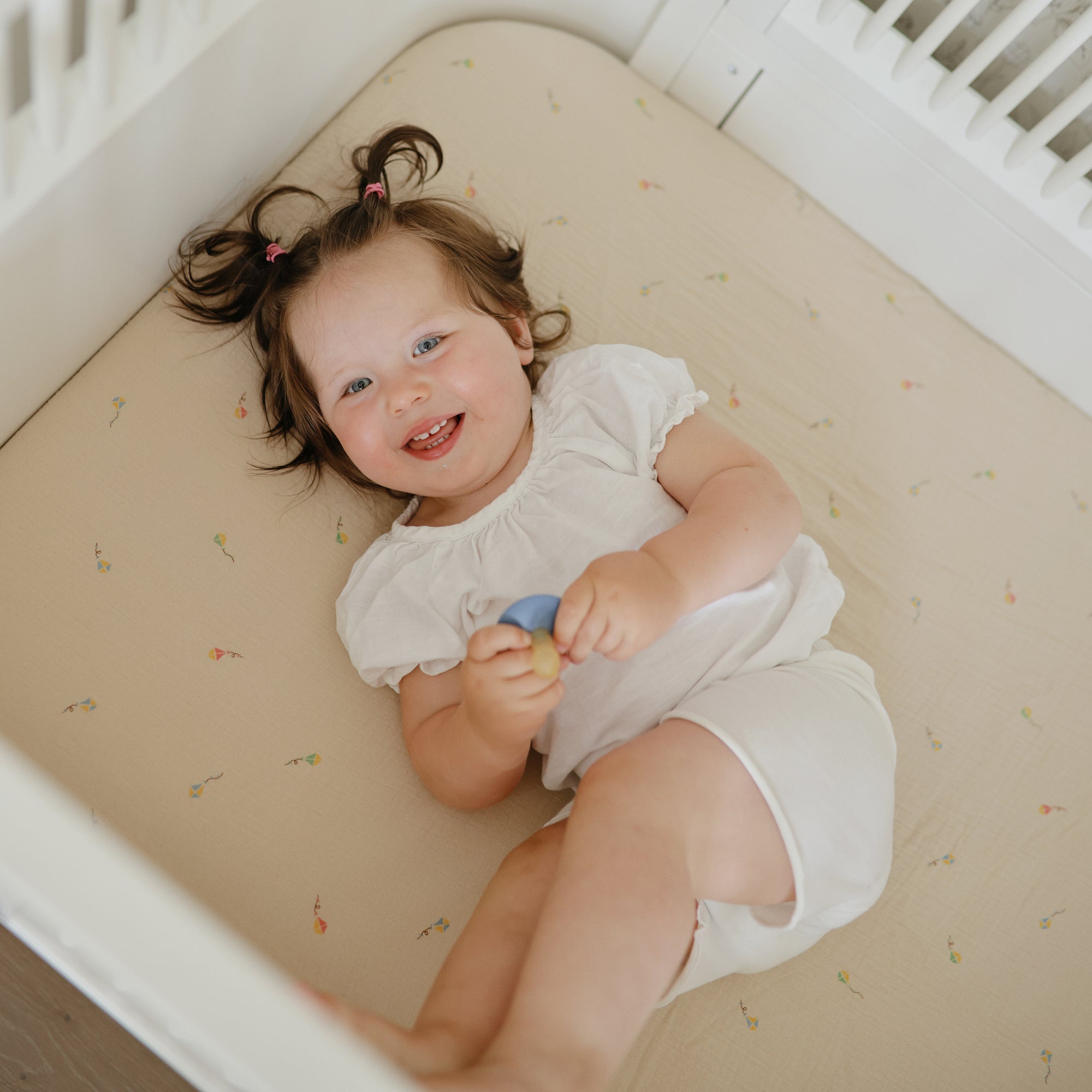 Happy toddler on mushie Extra Soft Muslin Crib Sheet, holding a toy and looking up with joy.