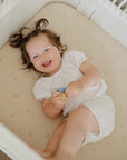 Happy toddler on mushie Extra Soft Muslin Crib Sheet, holding a toy and looking up with joy.