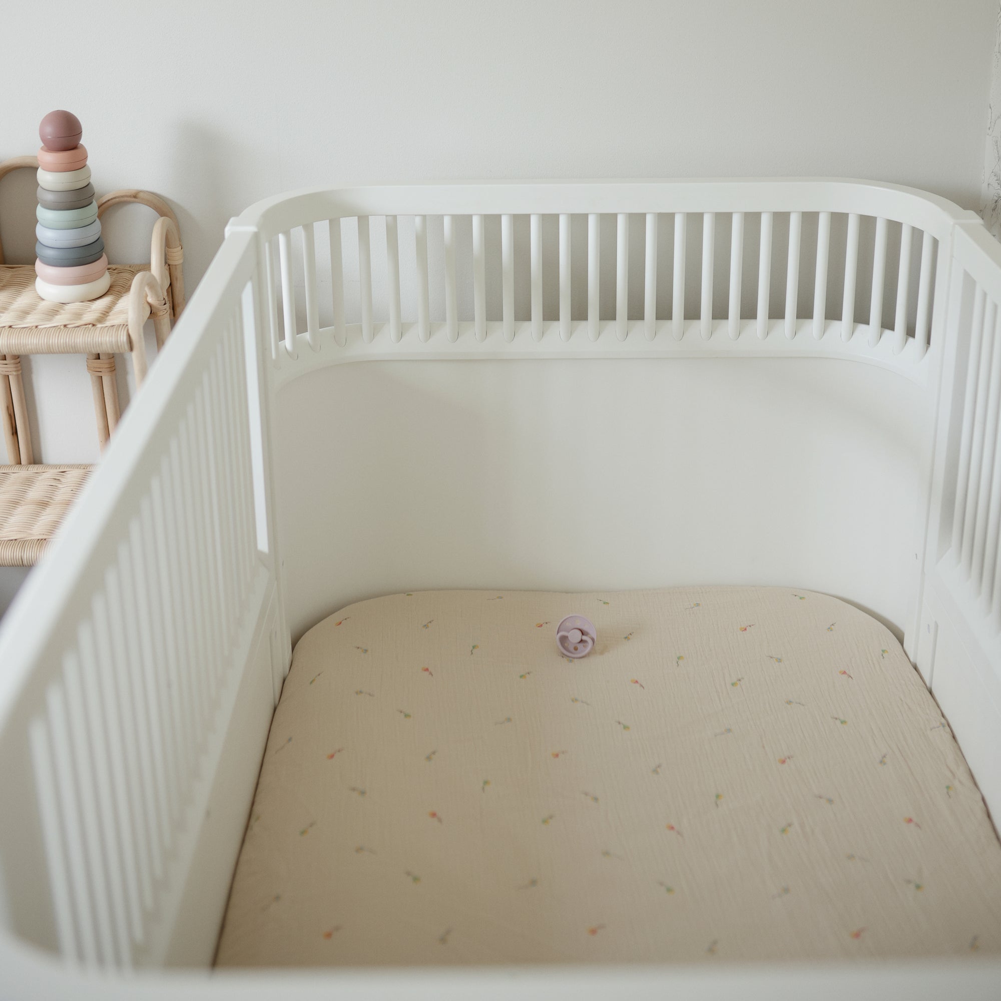 White crib with mushie Extra Soft Muslin Crib Sheet, purple pacifier, and stacked toy rings.