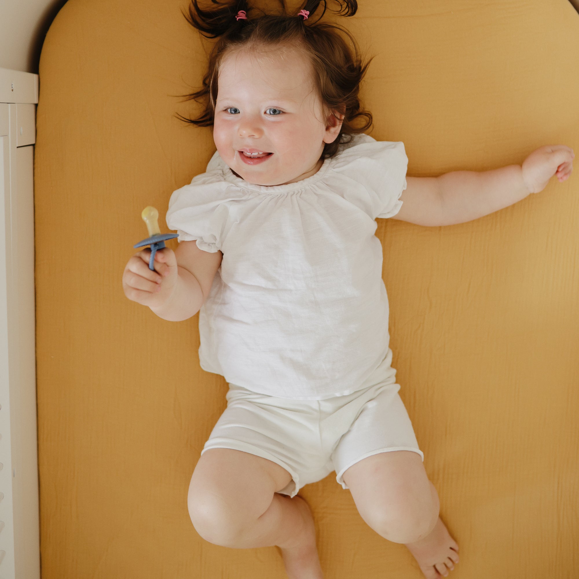 Smiling baby in white holds Mushie FRIGG Rope Natural Rubber Pacifier, lying on tan surface.
