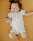 Smiling baby in white holds Mushie FRIGG Rope Natural Rubber Pacifier, lying on tan surface.