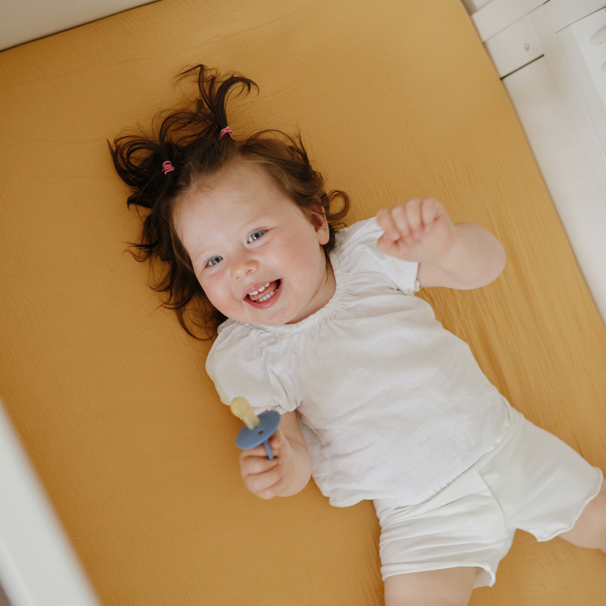 Smiling toddler on mushie Extra Soft Muslin Crib Sheet, holding pacifier, in white clothes.
