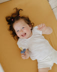 Smiling toddler on mushie Extra Soft Muslin Crib Sheet, holding pacifier, in white clothes.