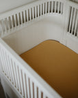 White baby crib with mushie Extra Soft Muslin Crib Sheet in yellow, placed in the corner.