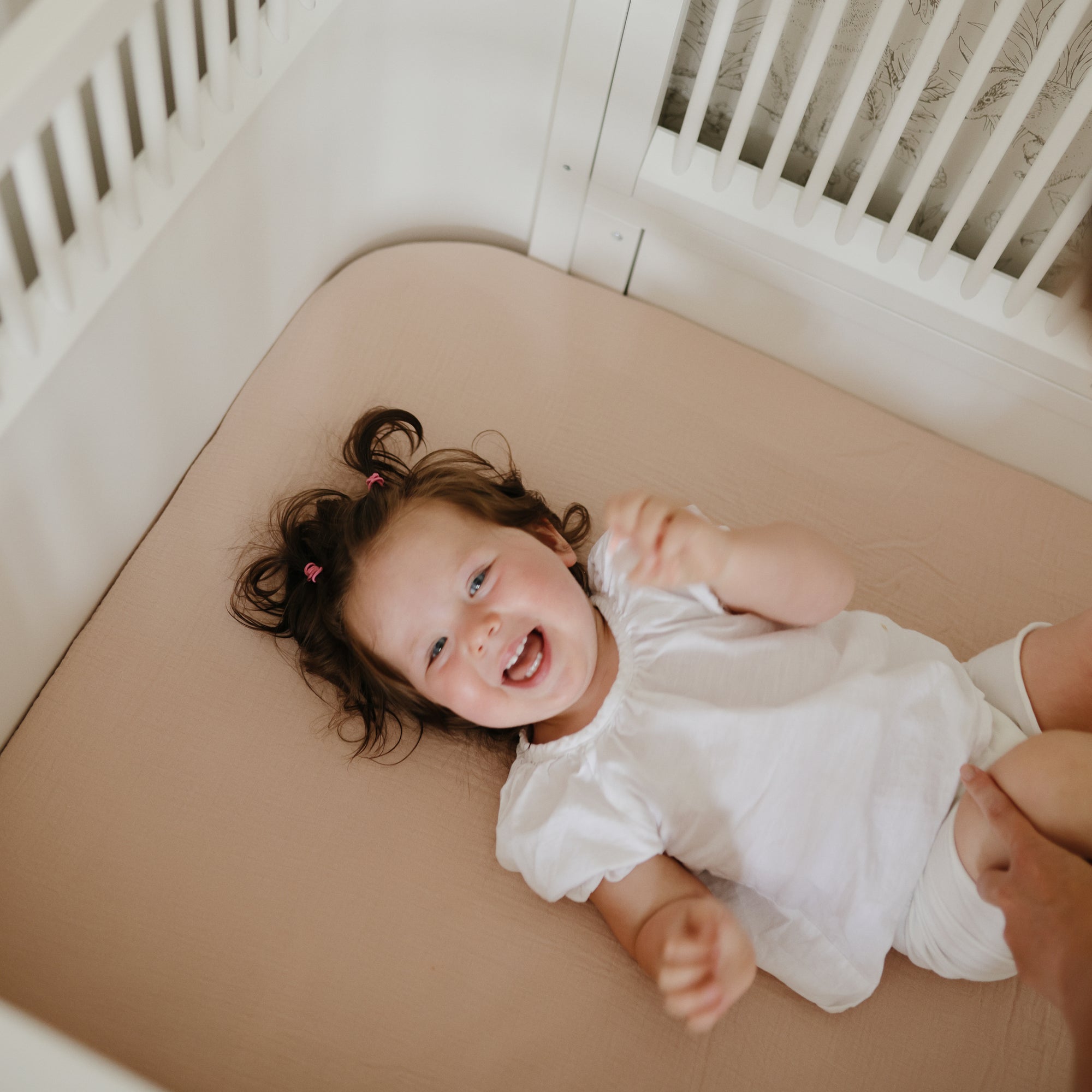 Happy toddler in white on mushie Extra Soft Muslin Crib Sheet in light brown cotton.