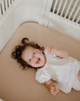 Happy toddler in white on mushie Extra Soft Muslin Crib Sheet in light brown cotton.