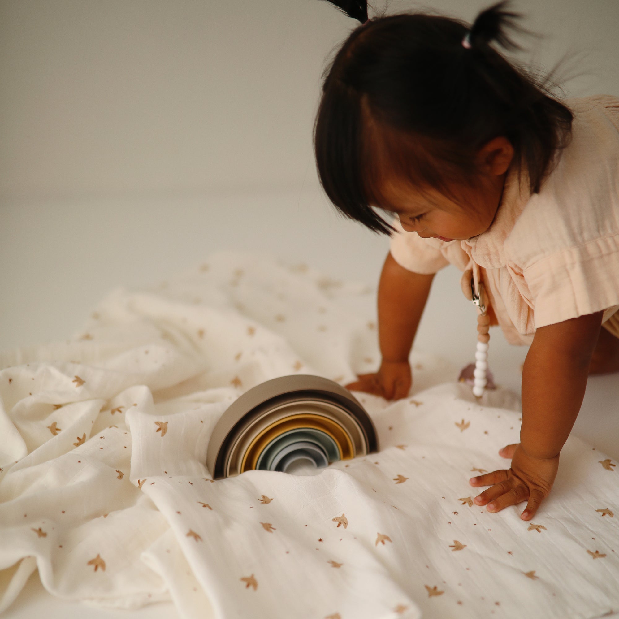 A toddler plays with stacking bowls on a mushie Organic Cotton Muslin Swaddle Blanket.
