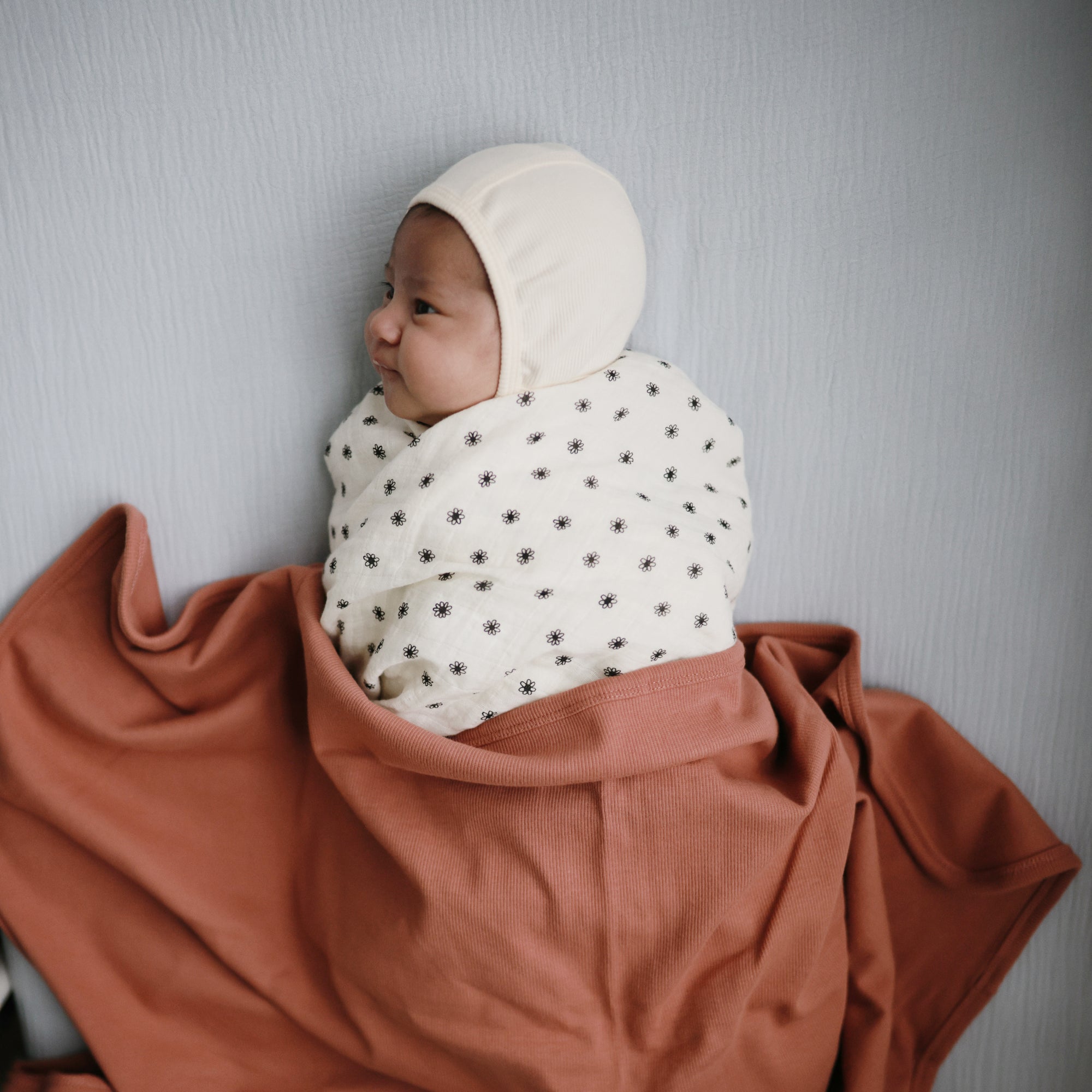 Baby swaddled in mushie Organic Cotton Muslin Swaddle Blanket on rust cloth on a gray surface.