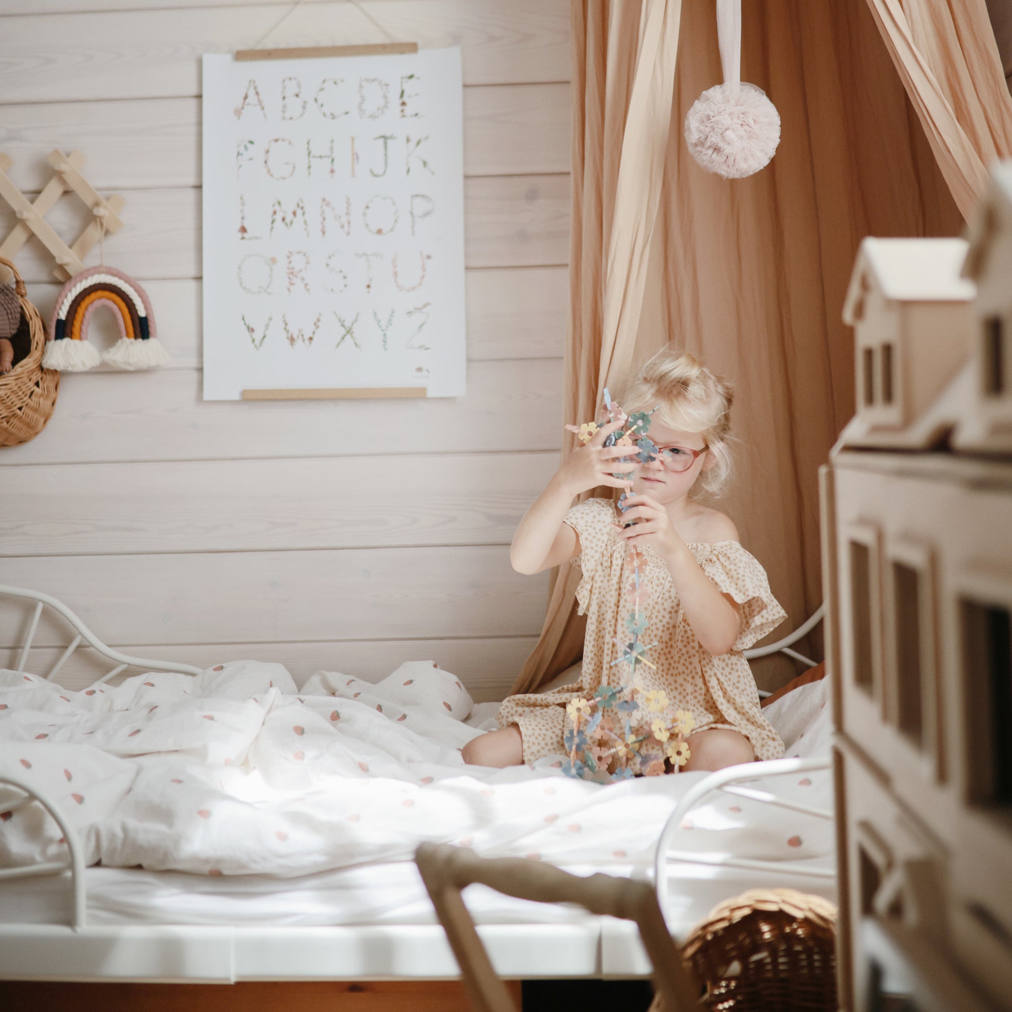 Young girl plays with mushie's HEX Connect Building Toy Set on a bed in a cozy bedroom.
