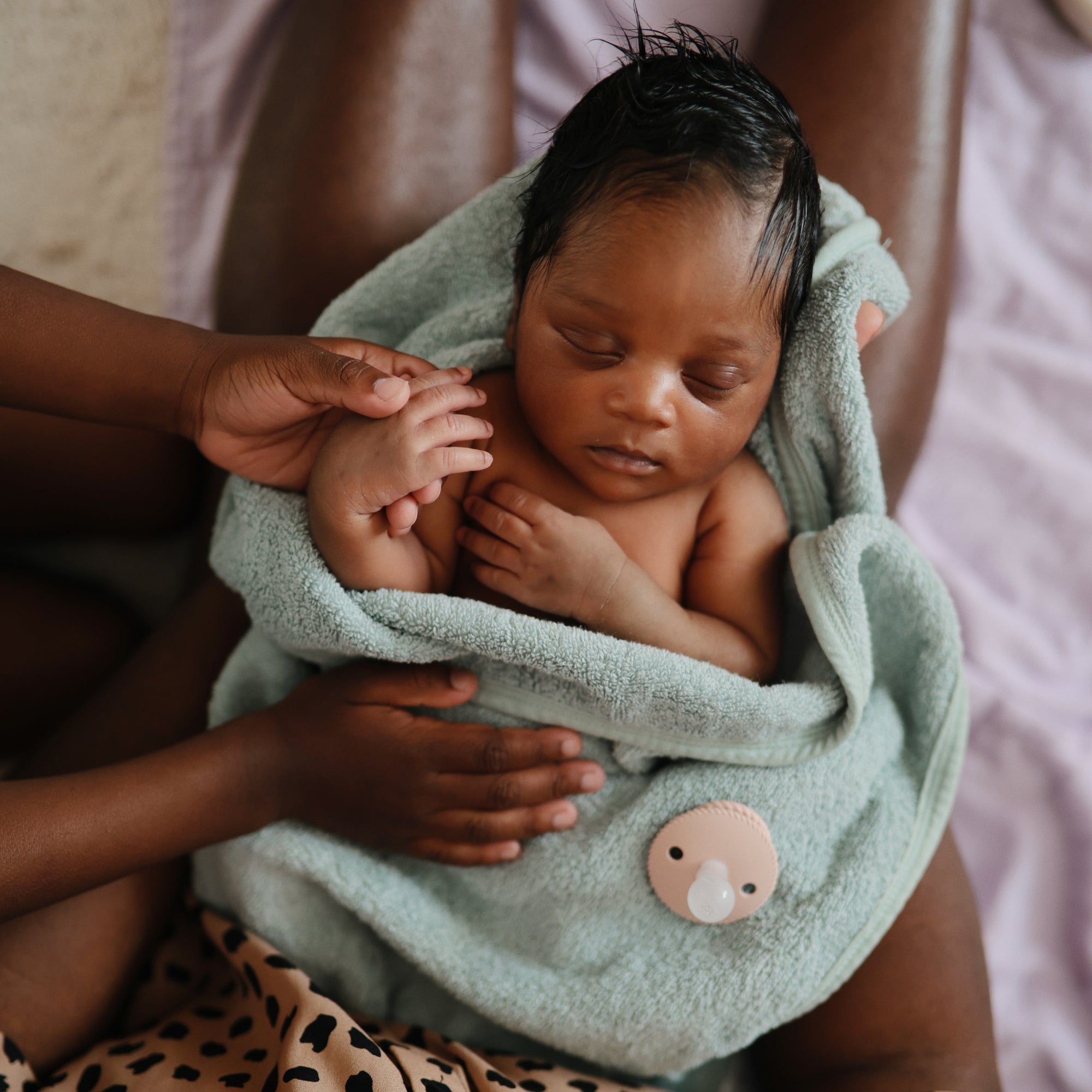 A newborn wrapped in a soft green towel rests on an adult’s lap, with a FRIGG Rope Silicone Pacifier 2-Pack by FRIGG nearby as an adult hand gently touches the baby’s arm—capturing the warmth of true baby essentials.