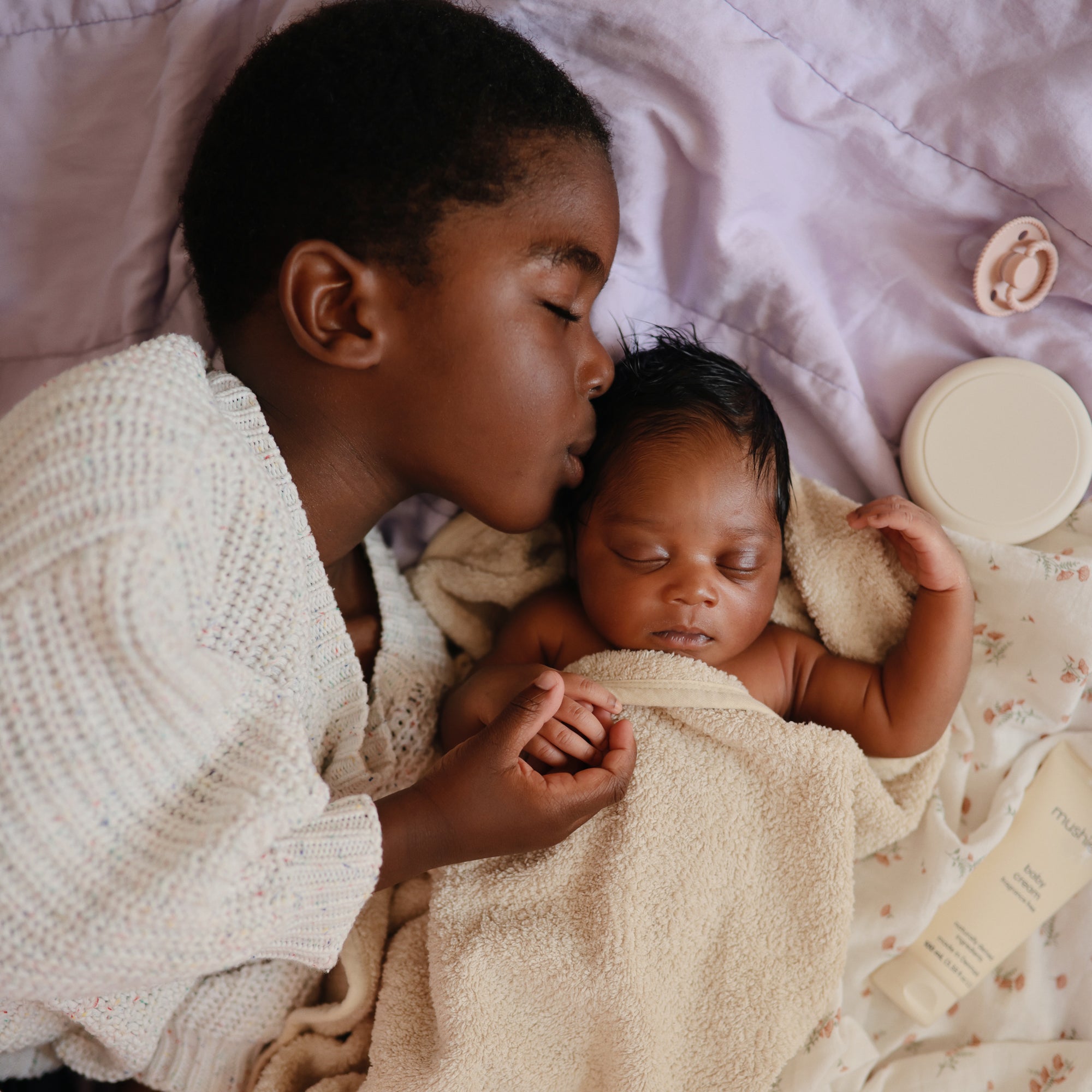 A child kisses a baby wrapped in a towel, their skin soft with mushie Baby Cream.