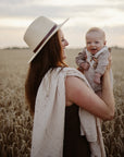Woman in a hat holds a baby wrapped in a mushie Organic Cotton Muslin Swaddle Blanket at sunset.