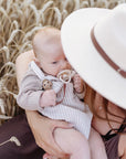 Woman in hat holds baby with mushie Linen Pacifier Clip 2-Pack in field of tall wheat grass.