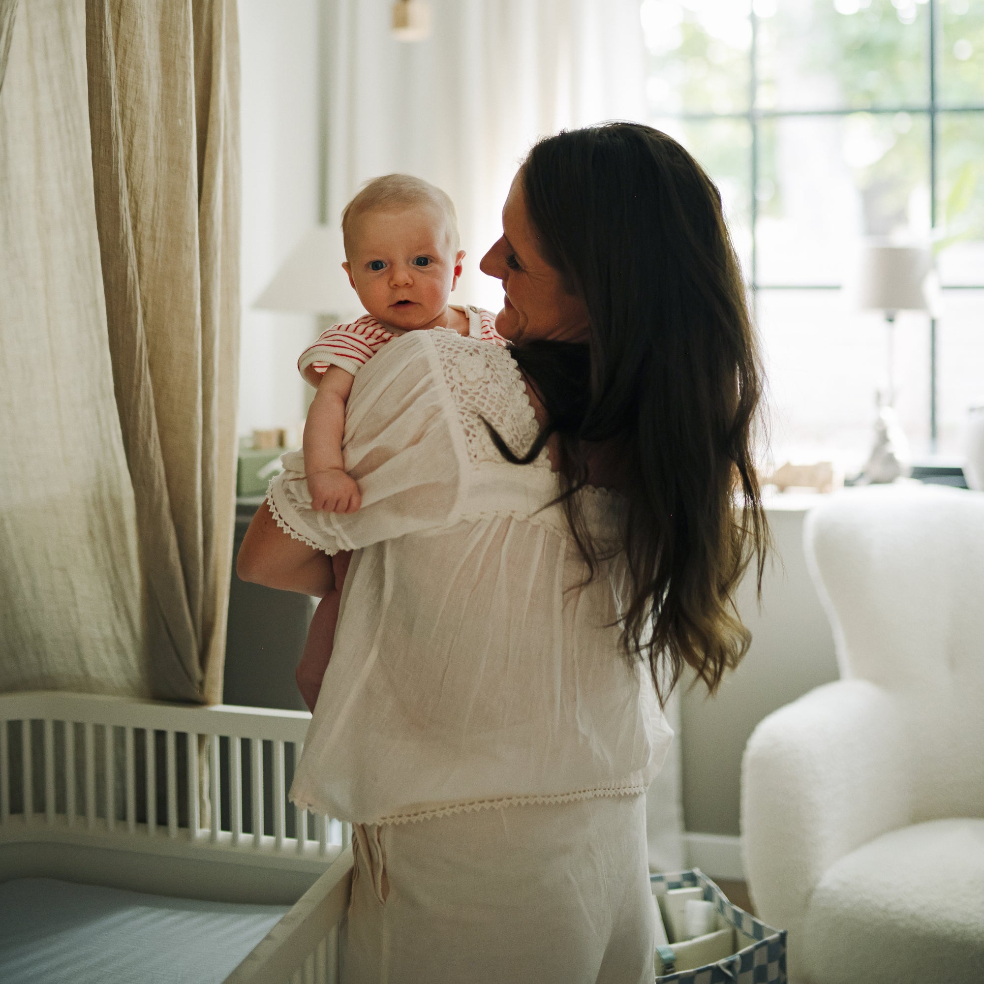 Woman holds baby in nursery with mushie Extra Soft Muslin Crib Sheet and breathable bedding.