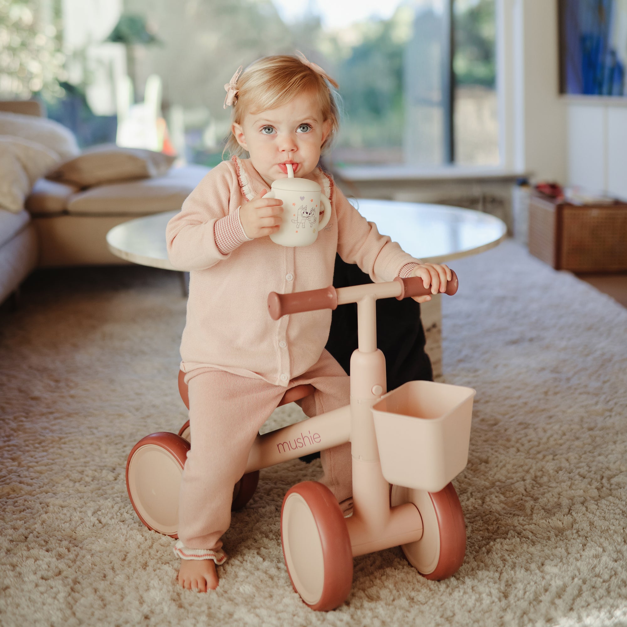 Toddler on a pink tricycle sipping from a Mushie Bluey x Mushie Silicone Training Cup + Straw.