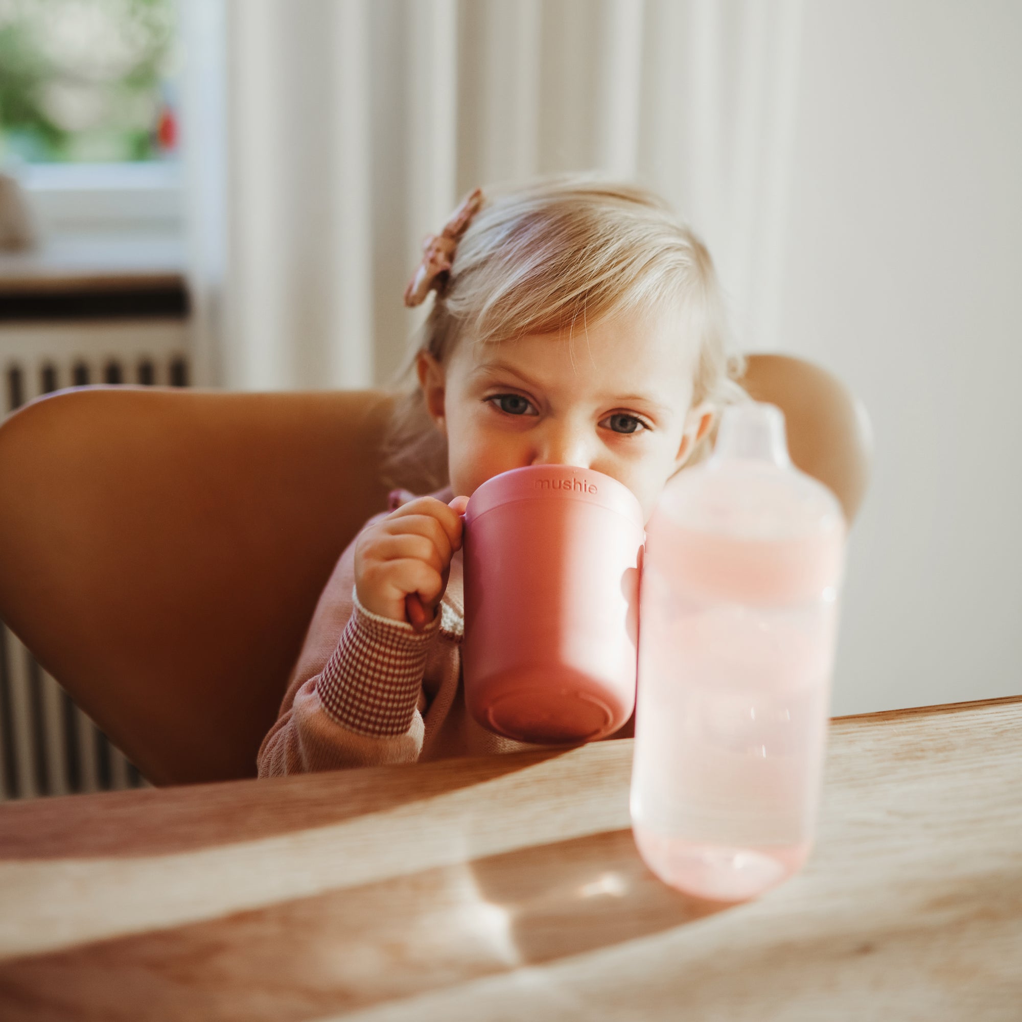 Young child uses Mushie Trainer Sippy Cup with easy-grip handles at the table.