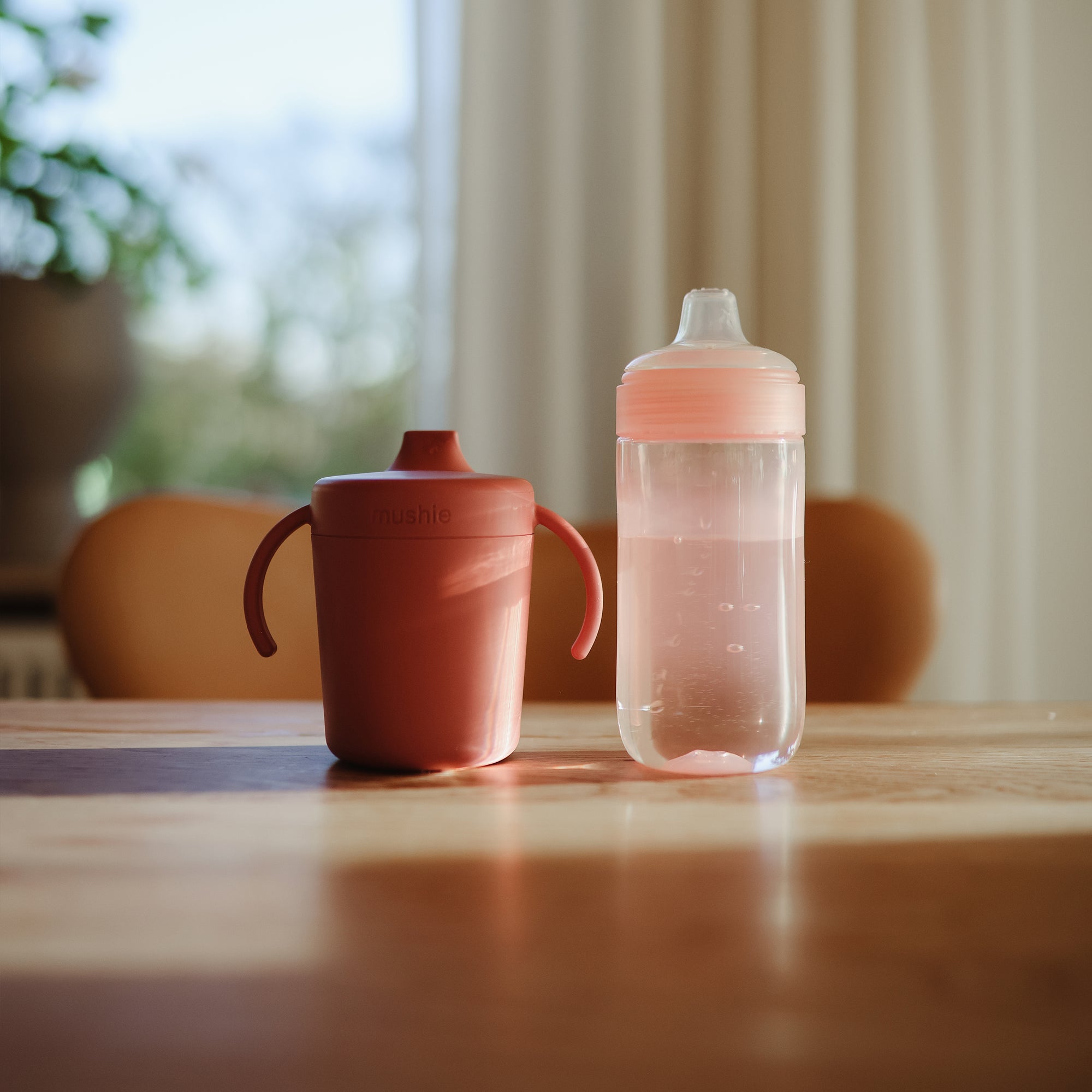 Two mushie Soft Spout Sippy Cups, including a pink one, sit on a sunlit table.