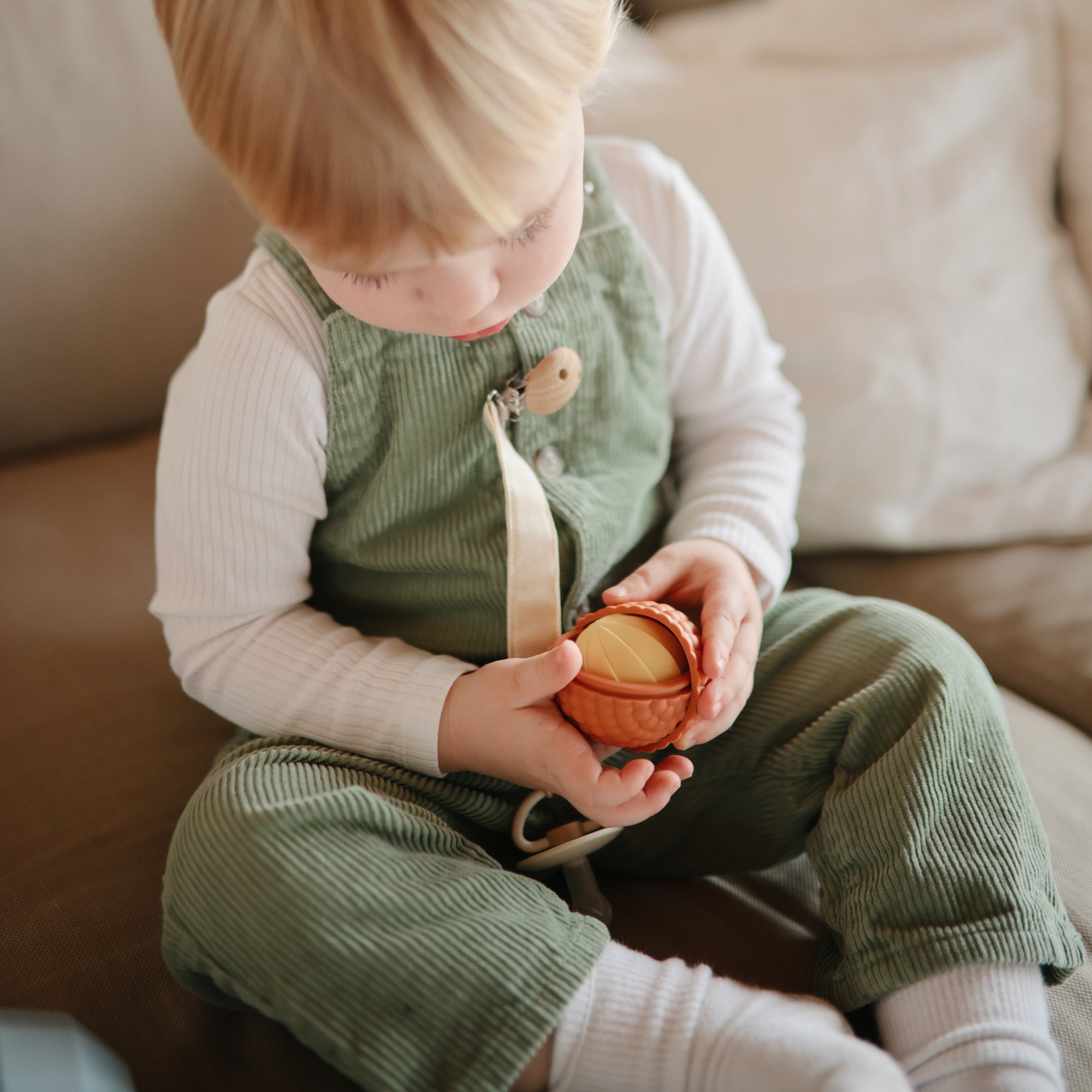Toddler in green overalls holds mushie Nesting Spheres Sensory Toy while sitting on the couch.