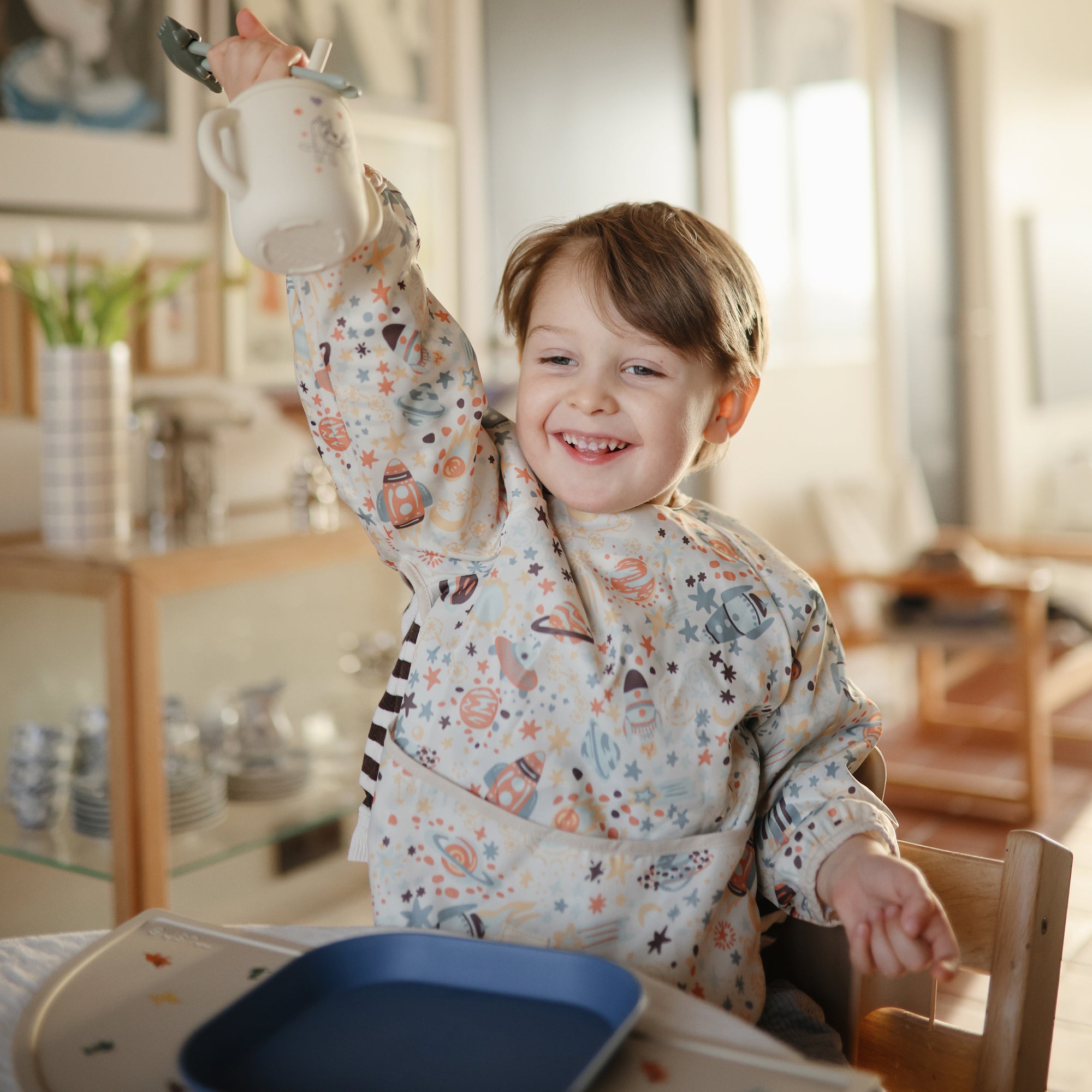 Smiling child holds a Mushie Bluey x Mushie Silicone Training Cup + Straw at the dining table.