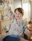 Smiling child holds a Mushie Bluey x Mushie Silicone Training Cup + Straw at the dining table.
