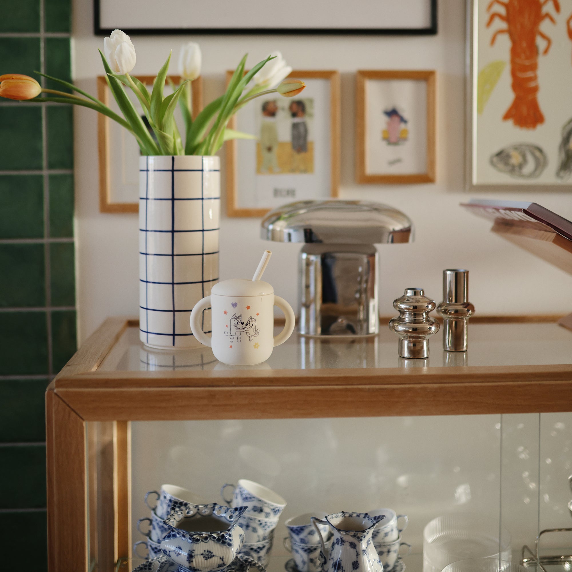 A glass display cabinet holds blue and white teacups, a vase of tulips, a Mushie Bluey x Mushie Silicone Training Cup + Straw, and metallic salt and pepper shakers. Framed art hangs on the wall behind.