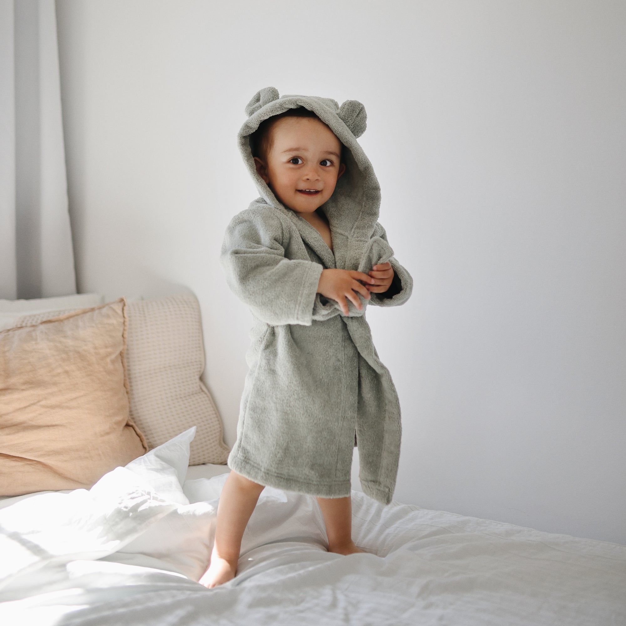 Toddlers stand on a bed wearing the mushie Bear Robe with hood and ears.