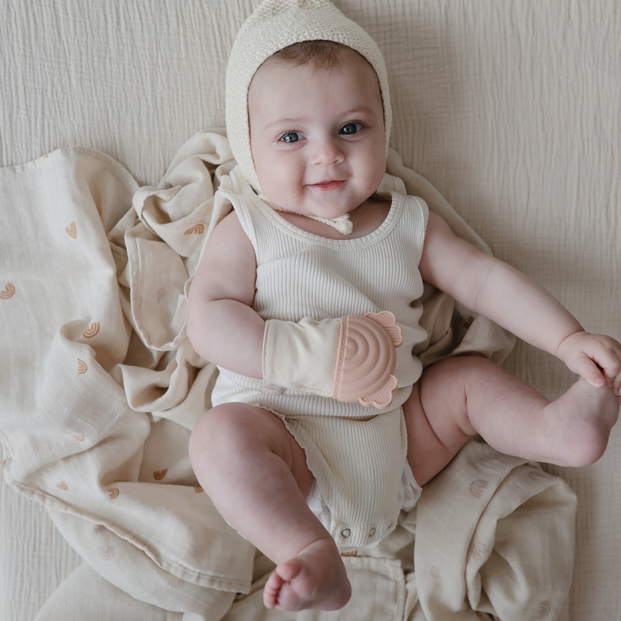 Smiling baby on mushie Organic Cotton Muslin Swaddle Blanket, wearing a teething mitten.