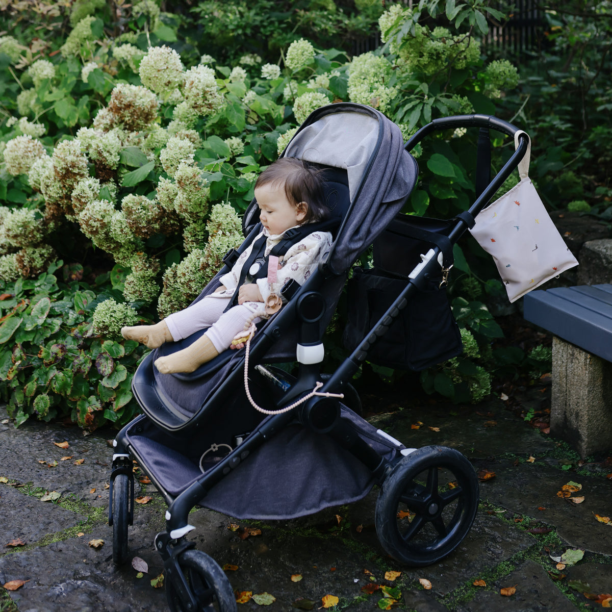 A toddler with a mushie Adjustable Silicone Strap 2-Pack sits in a gray stroller near green bushes.